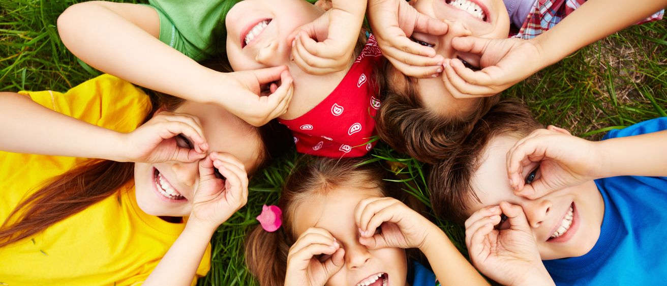 Mehrere Kinder liegen auf Gras und bilden einen Kreis, jeder mit den Händen auf den Augen, lächeln und lachen.