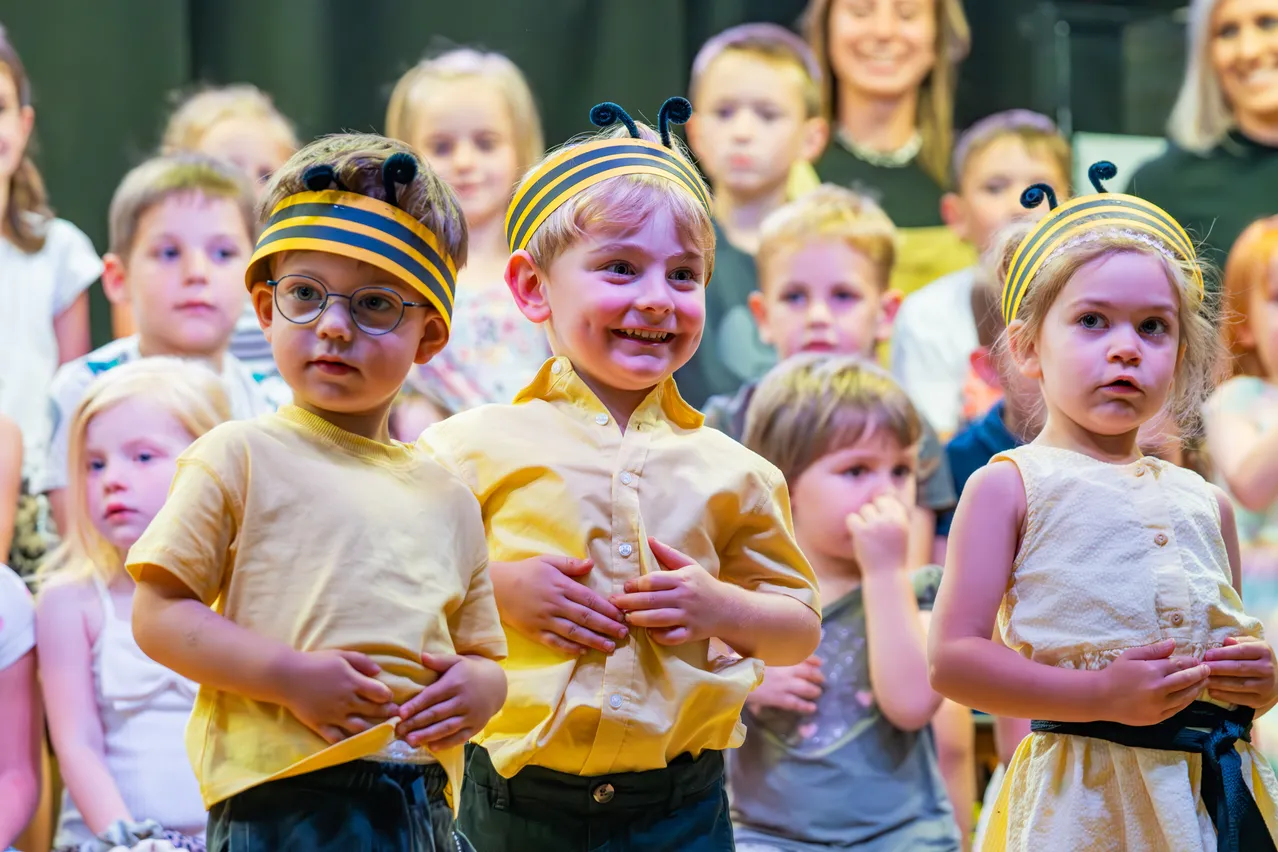 Zwei junge Jungen mit Bienenkopfbändern und gelben Hemden stehen in einem Klassenzimmer und lächeln. Hinter ihnen beobachtet eine vielfältige Gruppe von Kindern aufmerksam.