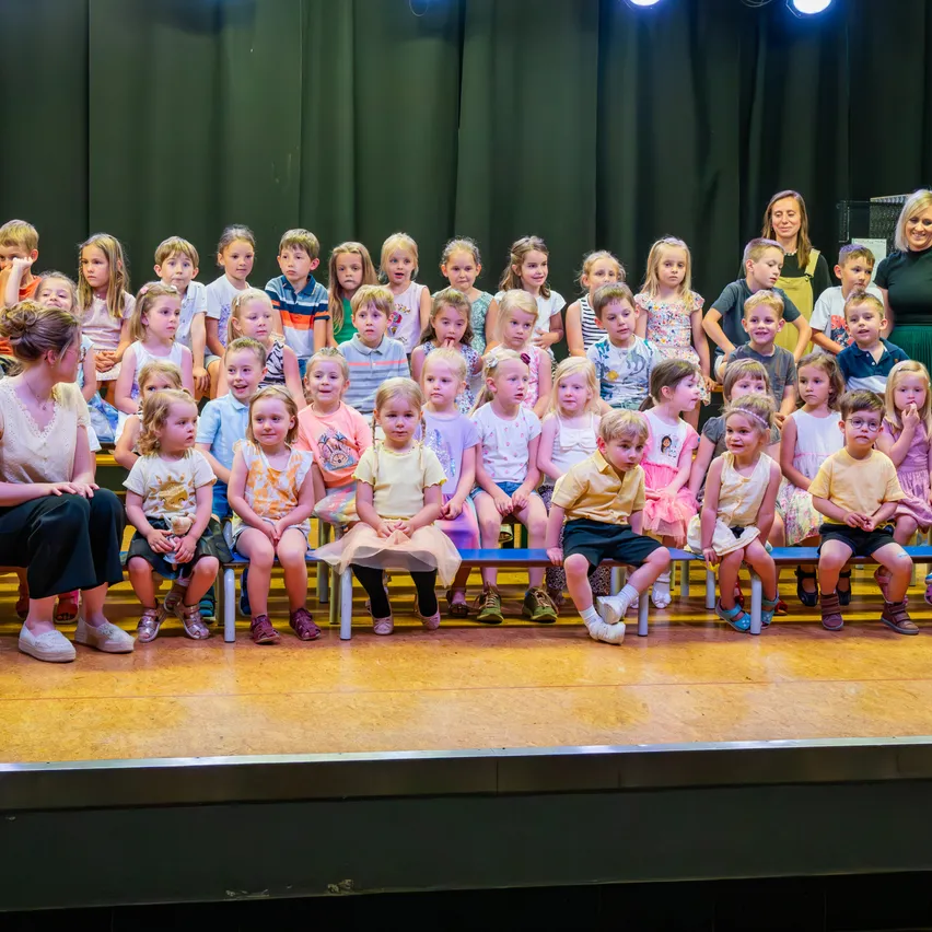 Eine Gruppe von Kindern sitzt auf der Bühne und trägt verschiedene Kleidungsstücke. Zwei Erwachsene stehen hinter ihnen. Die Bühne ist mit einem grünen Vorhang als Hintergrund gestaltet.