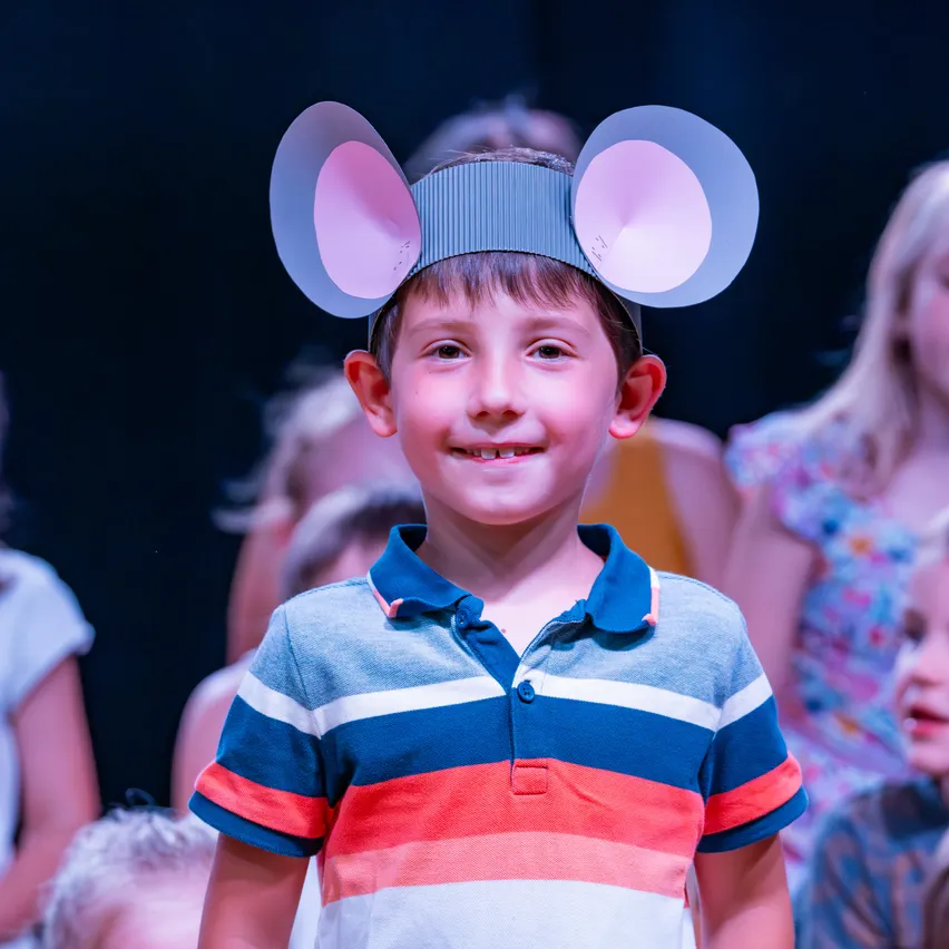 Ein junger Junge mit einem Papier-Maus-Kopfband lächelt in die Kamera, während andere im Hintergrund zusehen.