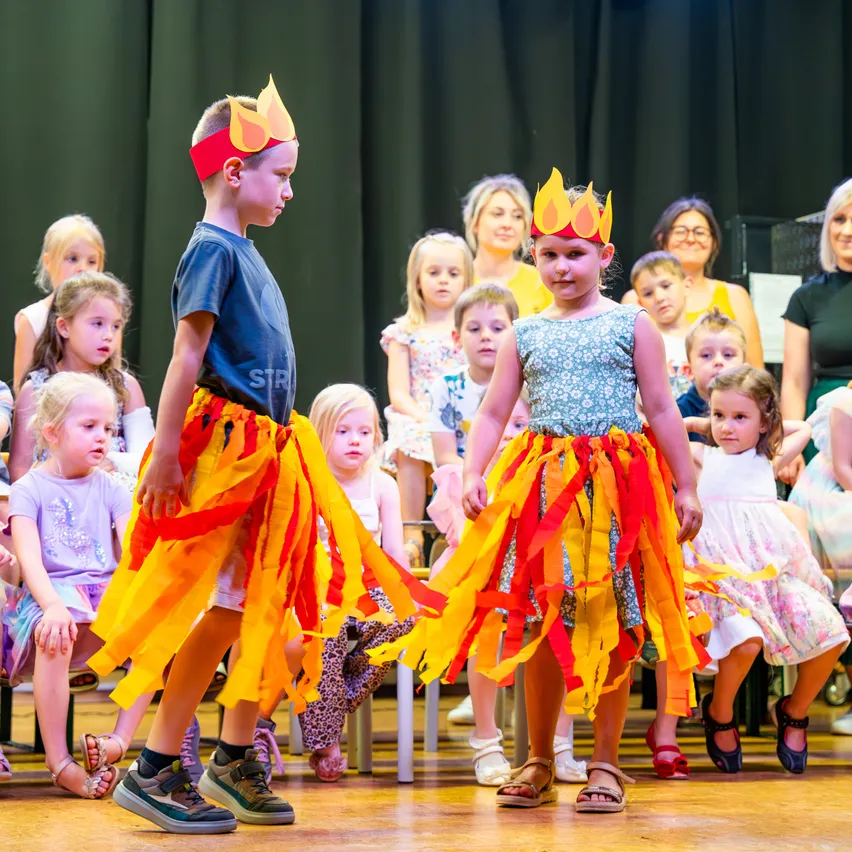 Zwei Kinder mit flammenförmigen Stirnbändern und bunten Röcken führen eine Aufführung auf der Bühne auf, umgeben von anderen Kindern und Erwachsenen, die zusehen.
