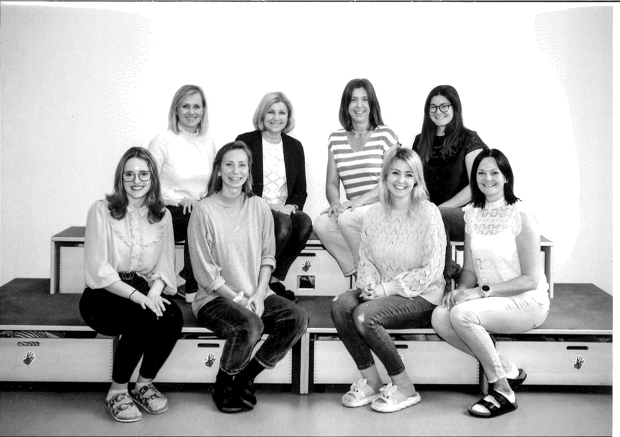 Eine Gruppe lächelnder Frauen sitzt auf einer Bank, gekleidet in verschiedene Outfits, in einem Schwarz-Weiß-Foto.