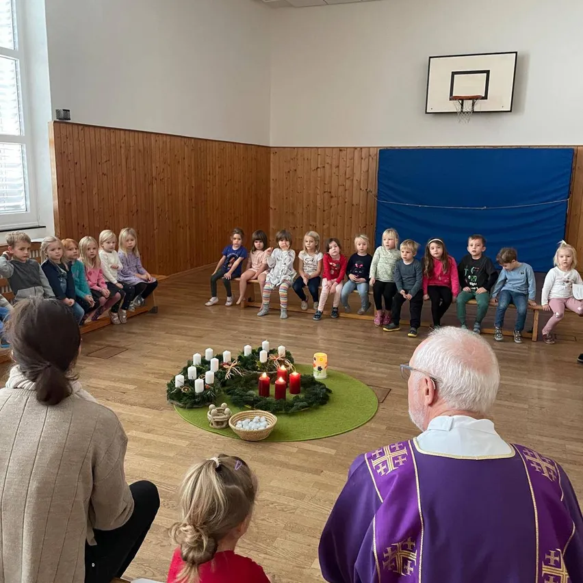 Eine Gruppe von Kindern in einer Kirche, die um einen Kranz mit Kerzen sitzen, während ein Priester und eine erwachsene Frau beobachten.