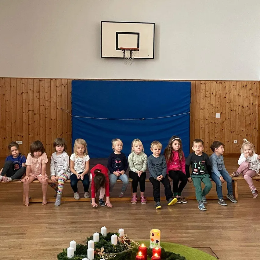 In einer Turnhalle sitzen mehrere Kinder auf einer Bank, vor ihnen eine Kerzenzeremonie. Die Turnhalle hat Holzwände und einen Basketballkorb.