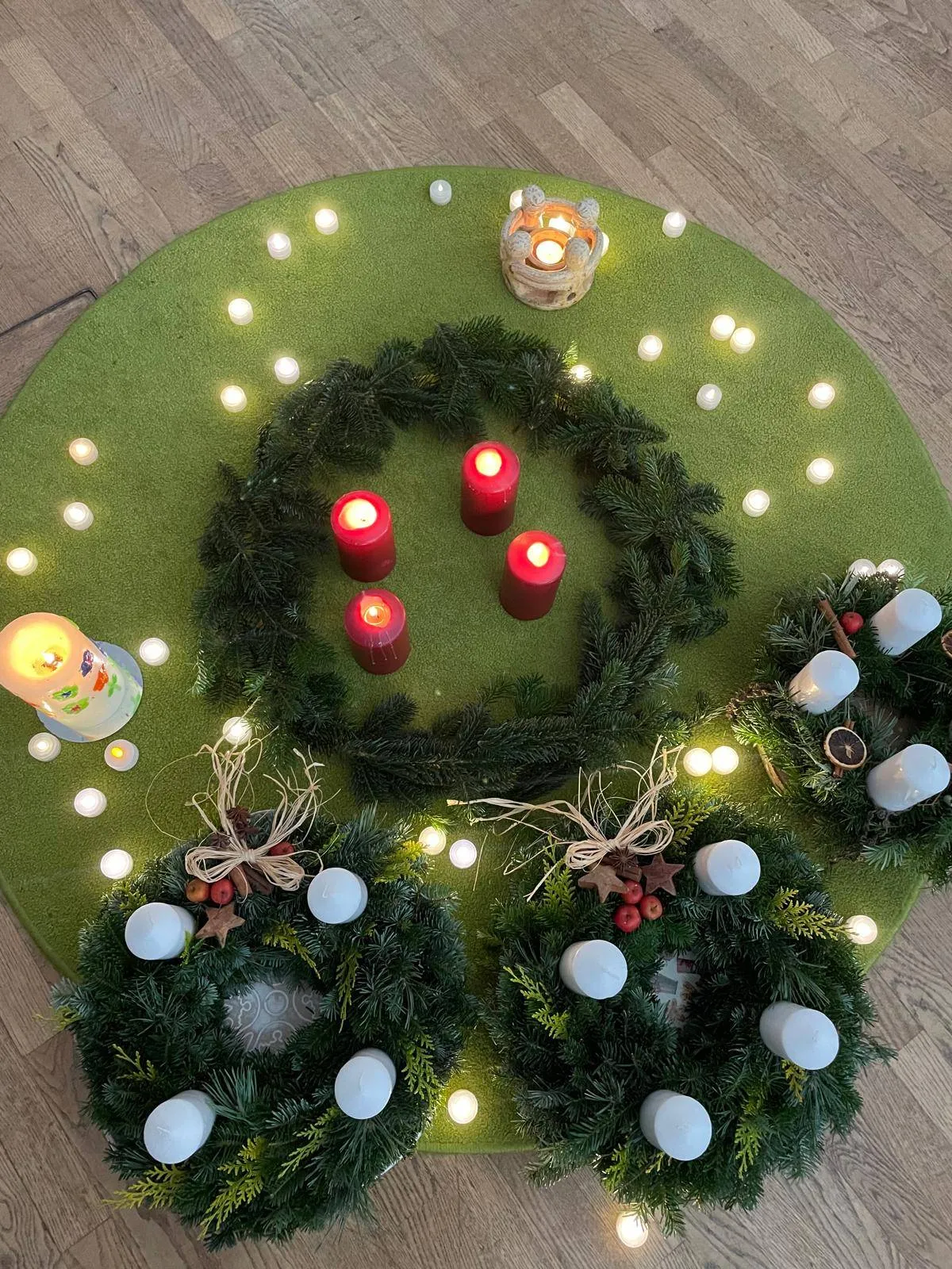 Ein grüner Kranz mit Kerzen darauf liegt auf einem grünen runden Teppich. Es gibt mehrere Kerzen auf dem Teppich und dem Kranz. Der Kranz hat ein Band und einige rote Beeren. Eine weiße Kerze ist auf der linken Seite platziert.