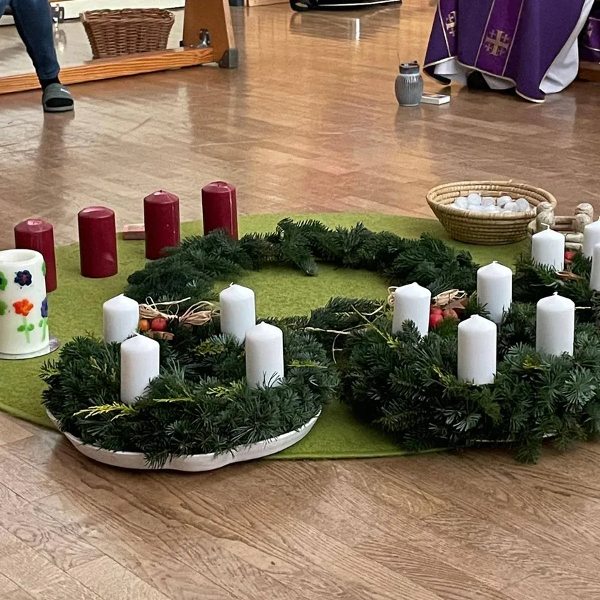 Ein Kranz aus grünen Tannenzweigen mit weißen Kerzen ist auf einem grünen Teppich angeordnet. Vier rote Kerzen sind auf der linken Seite platziert. Ein Korb mit Eiern und ein Tuch sind auf der rechten Seite platziert. Eine Person steht auf der linken Seite.