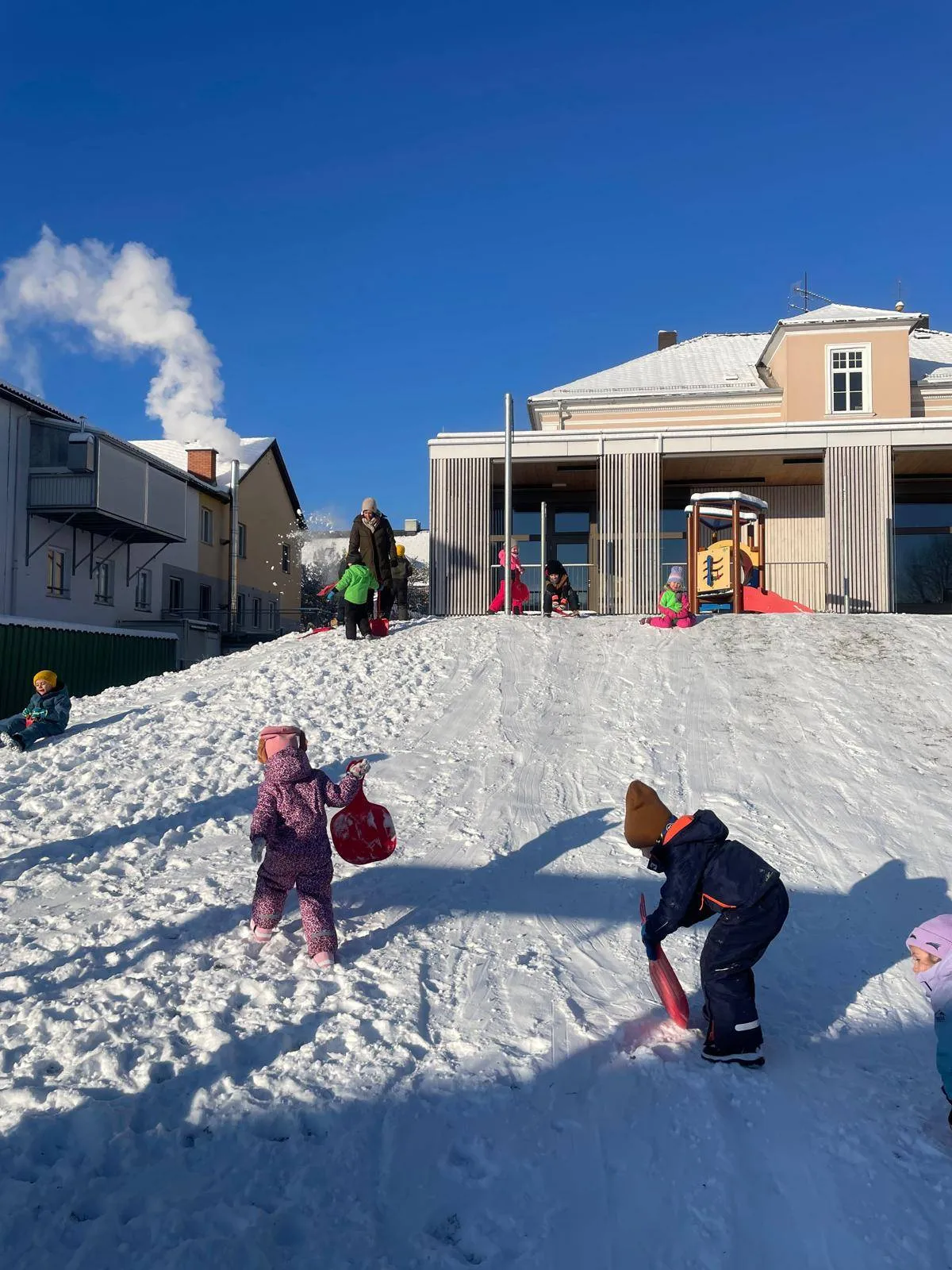 Kinder spielen auf einem verschneiten Hügel vor einem Gebäude. Einige Kinder sitzen im Schnee, während andere stehen und Schaufeln halten. Dahinter befindet sich ein Gebäude mit einem Schornstein, aus dem Rauch austritt.