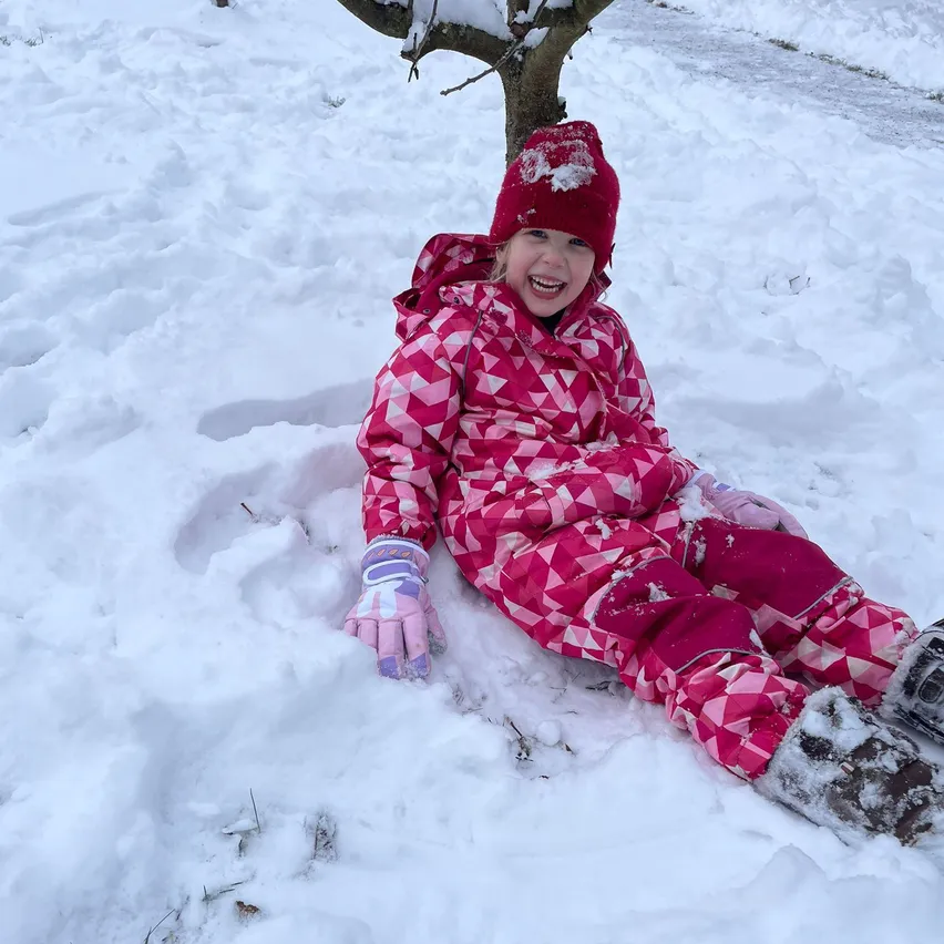 Ein kleines Mädchen in einem pinken Schneeanzug und Mütze sitzt im Schnee, lächelt und ist von Fußabdrücken umgeben. Schnee ist auf ihrem Kopf und ihren Stiefeln.
