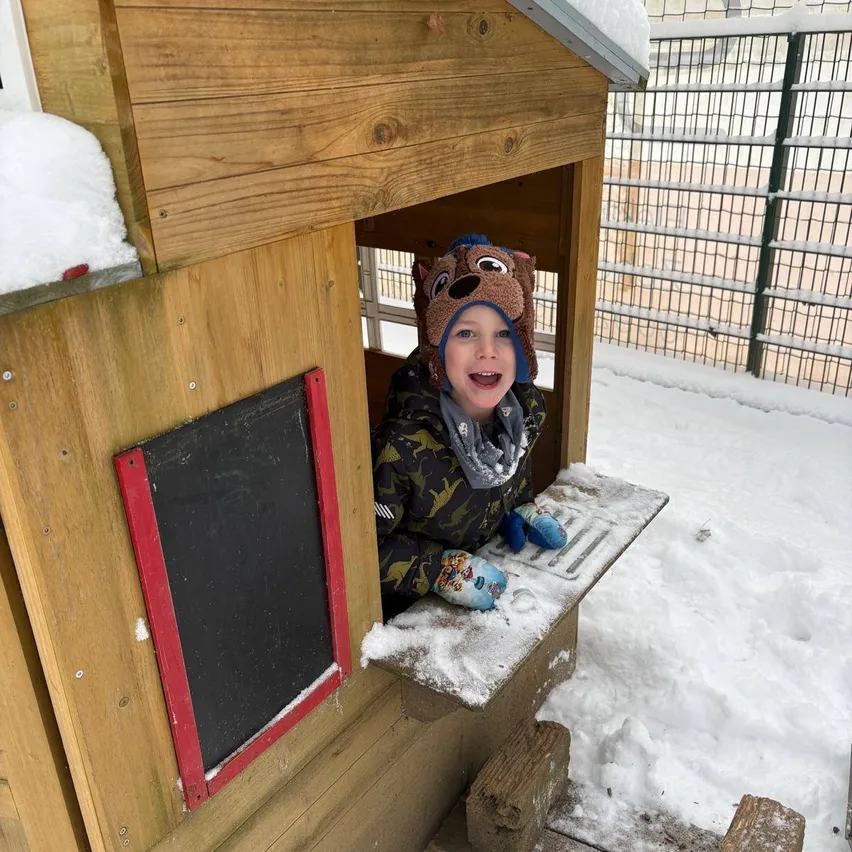 Ein Kind, das eine Wintermütze und Handschuhe trägt, sitzt in einem hölzernen Spielhaus und schaut mit einem Lächeln aus dem Fenster an einem verschneiten Tag.