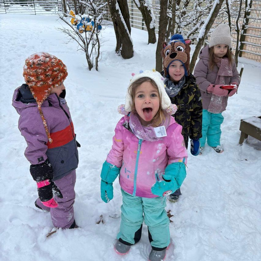 Vier Kinder stehen im Schnee. Ein Mädchen in Pink streckt die Zunge heraus, während die anderen Winterkleidung tragen.