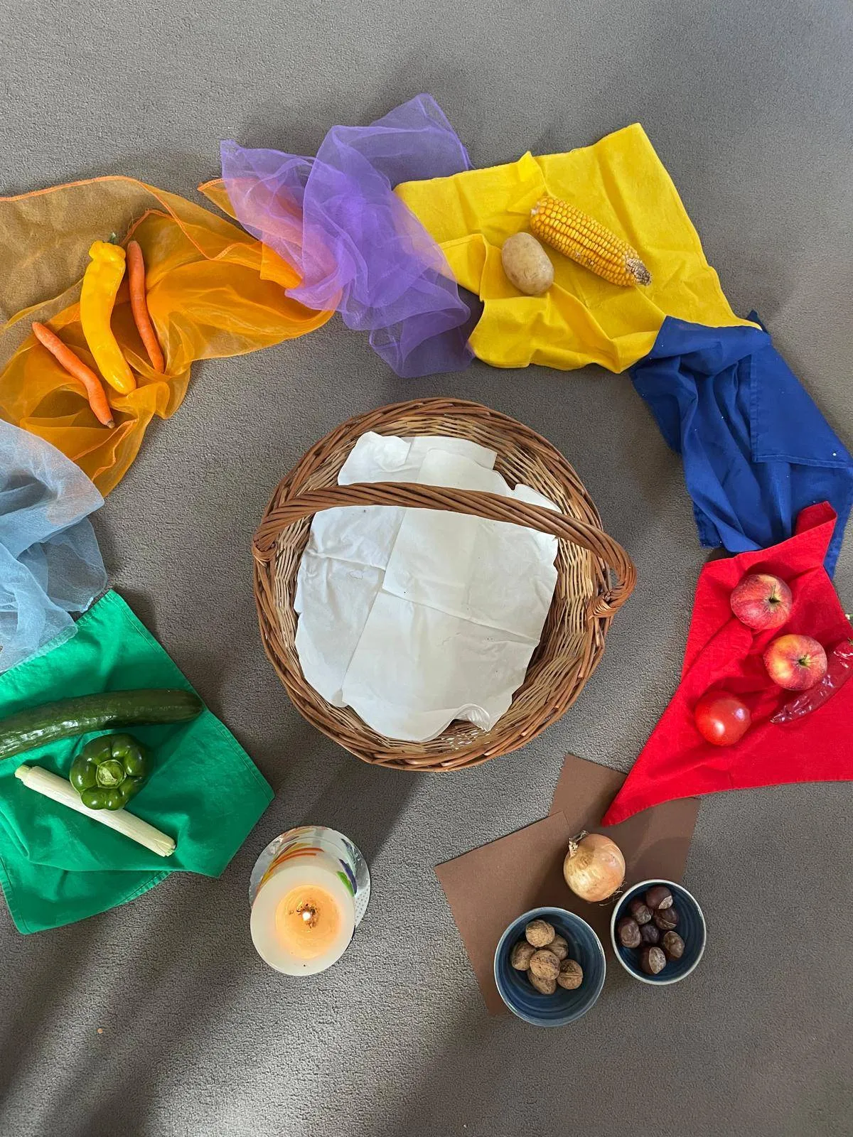 Ein Korb aus Weide mit weißem Papier liegt zwischen verschiedenen Gemüsesorten und farbigen Tüchern auf einem Teppichboden. In der Nähe befinden sich eine Kerze, Äpfel, Nüsse und Zwiebeln.