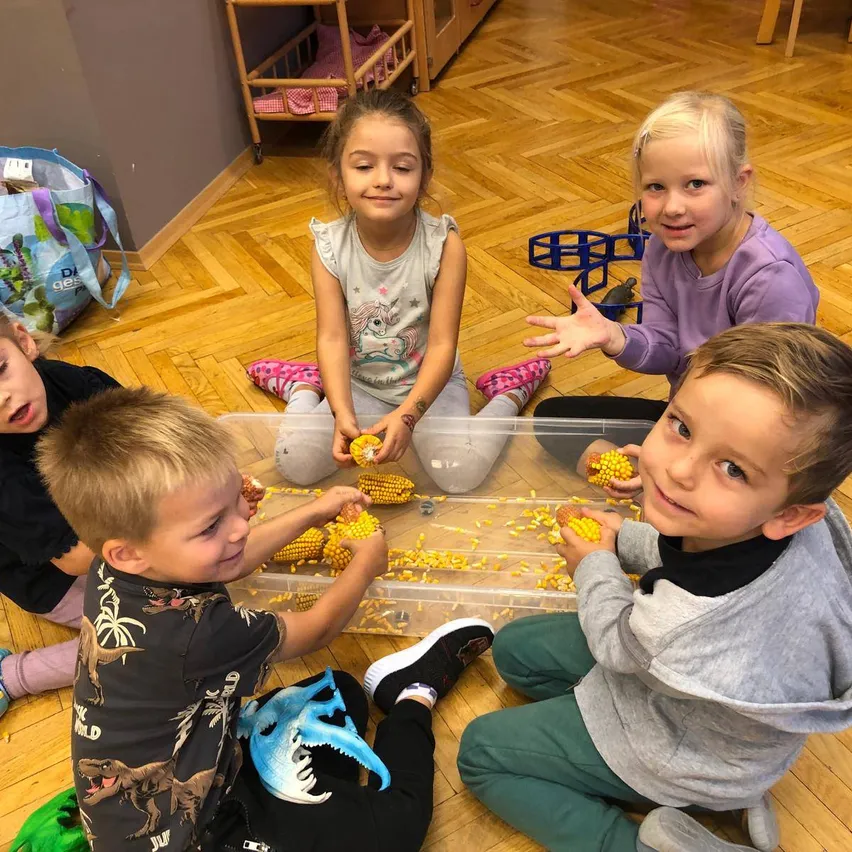 Fünf Kinder sitzen in einem Klassenzimmer auf dem Boden, ein Kind hält einen Behälter mit Mais. Sie lächeln und schauen den Mais an.