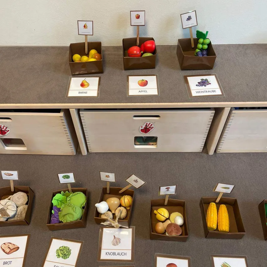 Eine Ausstellung verschiedener Obst- und Gemüsesorten in beschrifteten Kartonboxen auf einer grauen Oberfläche, mit zusätzlichen Schubladen darunter.