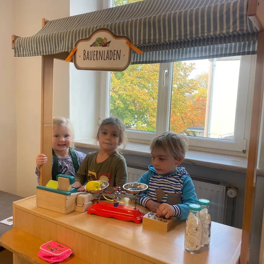 Drei Kinder spielen an einem kleinen hölzernen Ladentresen. Ein Schild darüber lautet 'Bauernladen'. Der Tresen ist mit einer Waage, Gläsern und einer Box ausgestattet. Sie sitzen vor einem Fenster mit Bäumen draußen.
