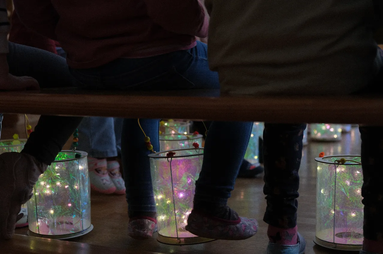 Mehrere Kinder sitzen mit leuchtenden Laternen auf dem Schoß in einem schwach beleuchteten Raum. Ihre Schuhe sind mit Lichtern verziert.