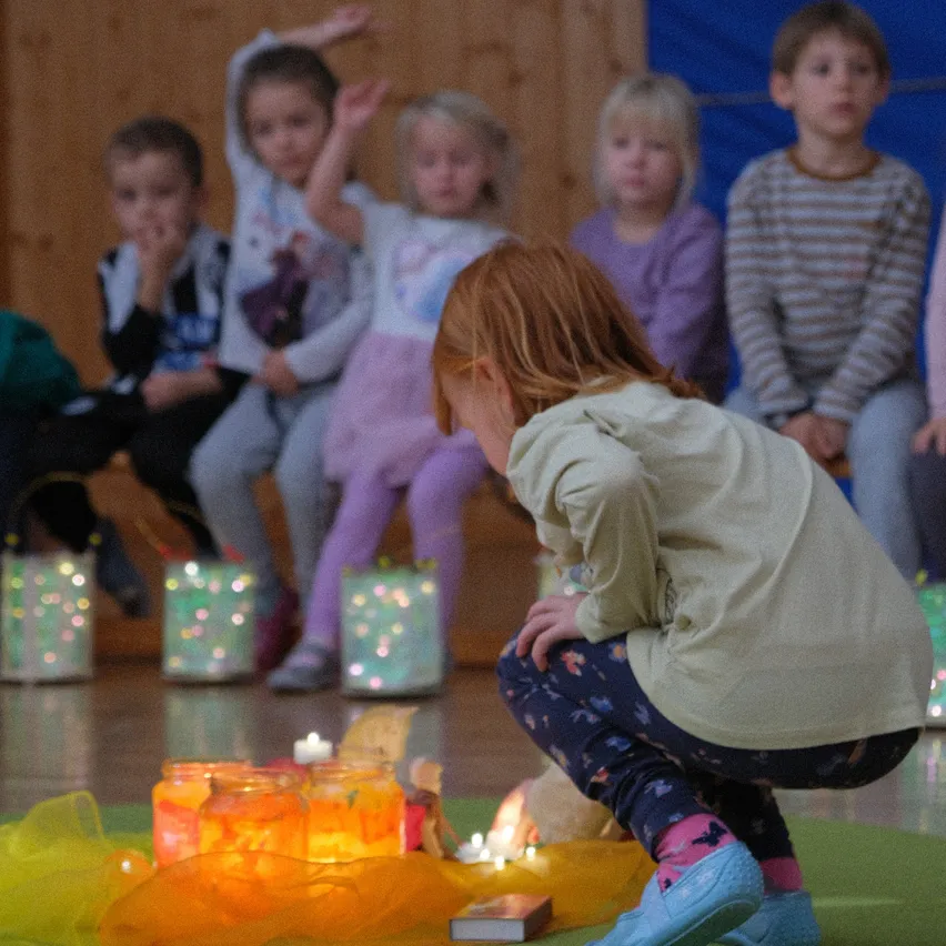 Kinder sitzen auf Bänken und beobachten ein Mädchen, das Kerzen vor ihnen anzündet. Sie trägt hellblaue Hausschuhe und eine weiße Kapuzenjacke. Kerzen stehen vor ihnen.