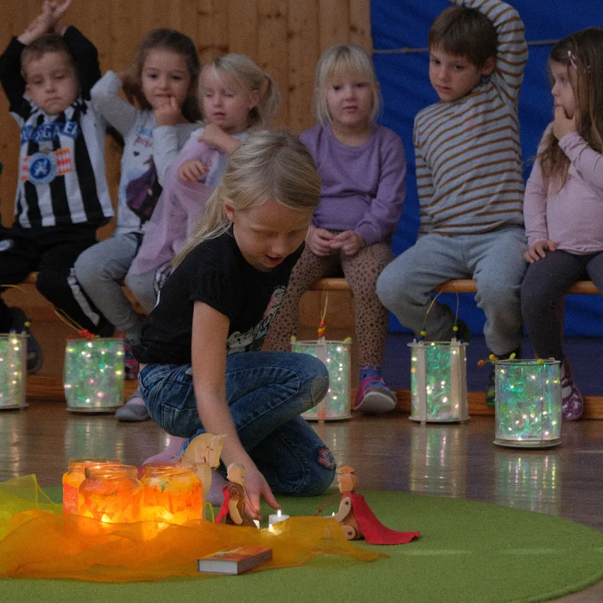 Eine Gruppe von Kindern, die auf einer Bank sitzen und einer jungen Mädchen zusehen, die mit Spielzeug und brennenden Kerzen auf einem grünen Teppich spielt.
