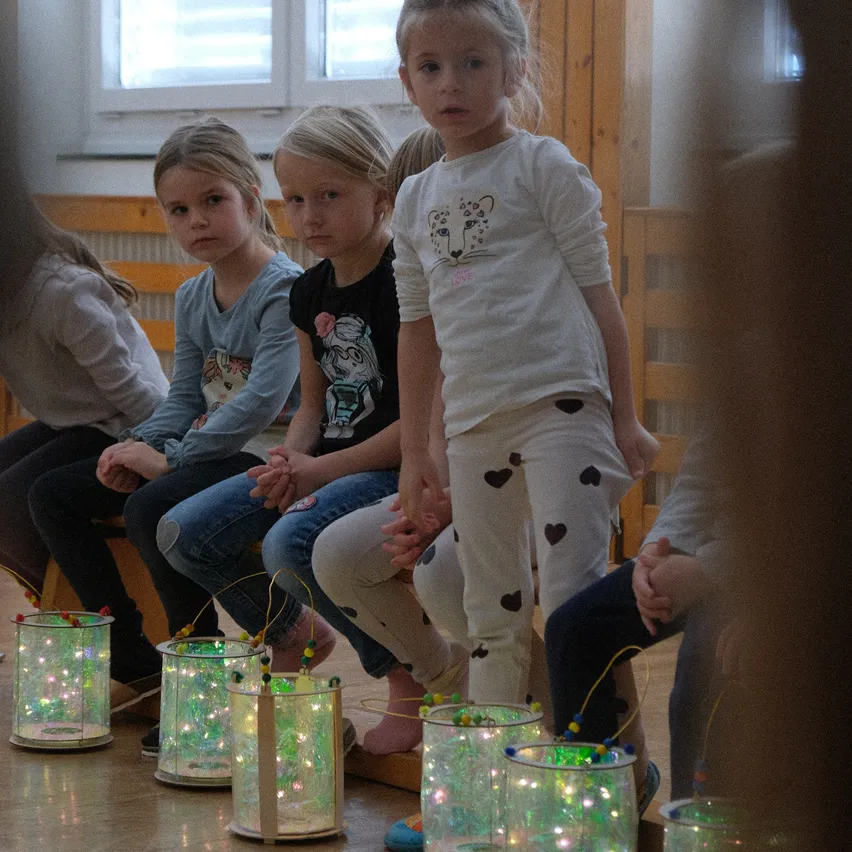 Vier junge Mädchen sitzen in einer Reihe, jede hält eine beleuchtete Laterne mit bunten Lichtern. Sie schauen nach vorne und nehmen wahrscheinlich an einer Klassenaktivität teil. Dahinter sitzen weitere Kinder.