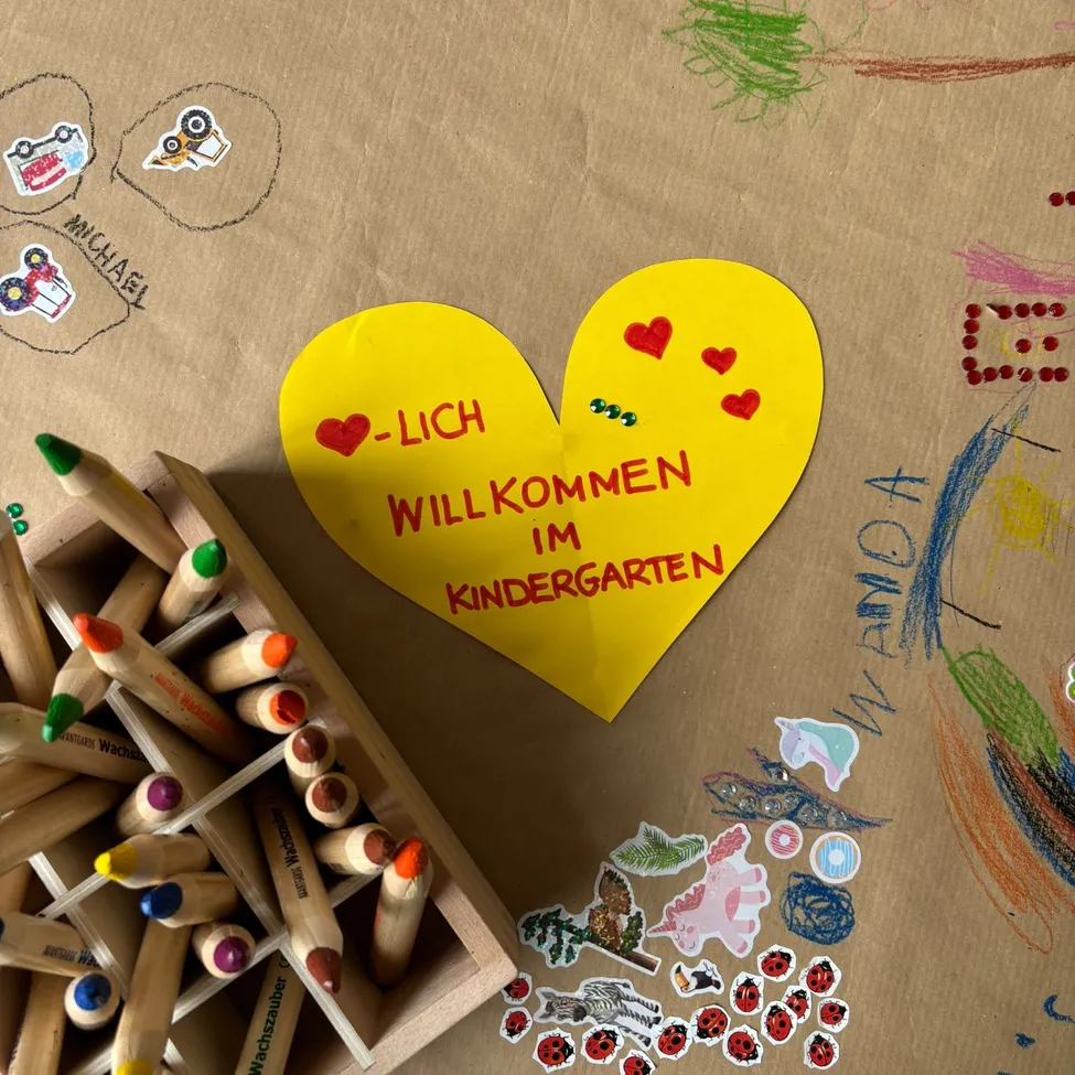 Ein gelbes Herz mit den Worten 'LICH WILLKOMMEN IM KINDERGARTEN' liegt auf einem braunen Papierhintergrund mit verschiedenen farbigen Stiften, Aufklebern und Zeichnungen.