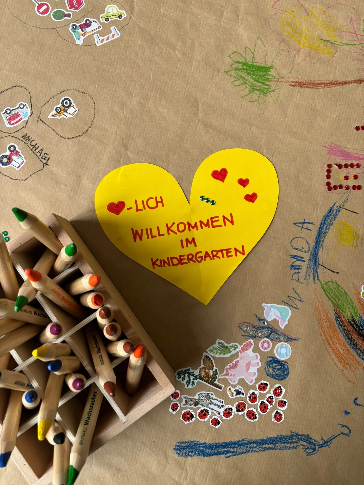 Ein gelbes Herz mit den Worten 'LICH WILLKOMMEN IM KINDERGARTEN' liegt auf einem braunen Papierhintergrund mit verschiedenen farbigen Stiften, Aufklebern und Zeichnungen.