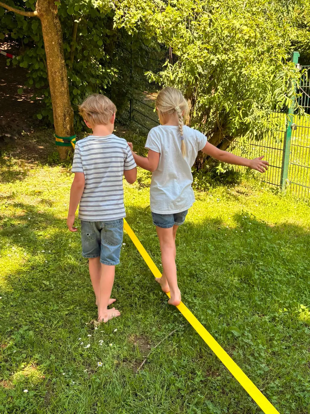 Zwei Kinder halten sich an den Händen und gehen auf einer gelben Linie auf dem Rasen. Dahinter befindet sich ein Baumstamm und ein grüner Zaun.