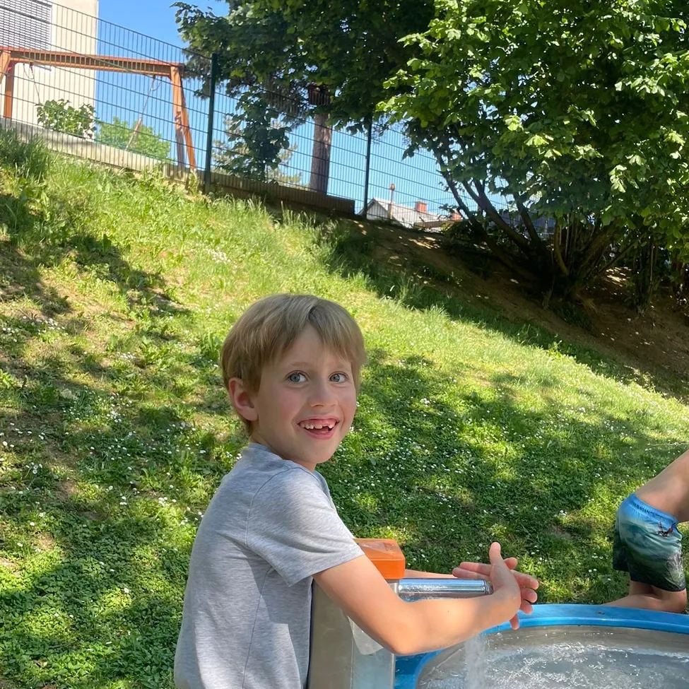 Ein kleiner Junge steht an einem Wasserbrunnen im Garten, lächelt und hält eine Seifenspender in der Hand. Ein weiteres Kind ist im Hintergrund in der Nähe eines hölzernen Schaukelgestells.