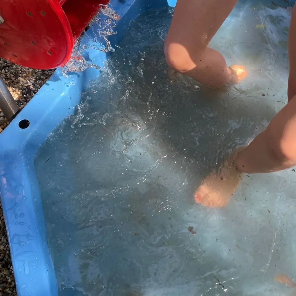 Eine Person steht in einem blauen aufblasbaren Pool, wobei Wasser um ihre Beine spritzt. Ein roter Eimer gießt Wasser in den Pool.
