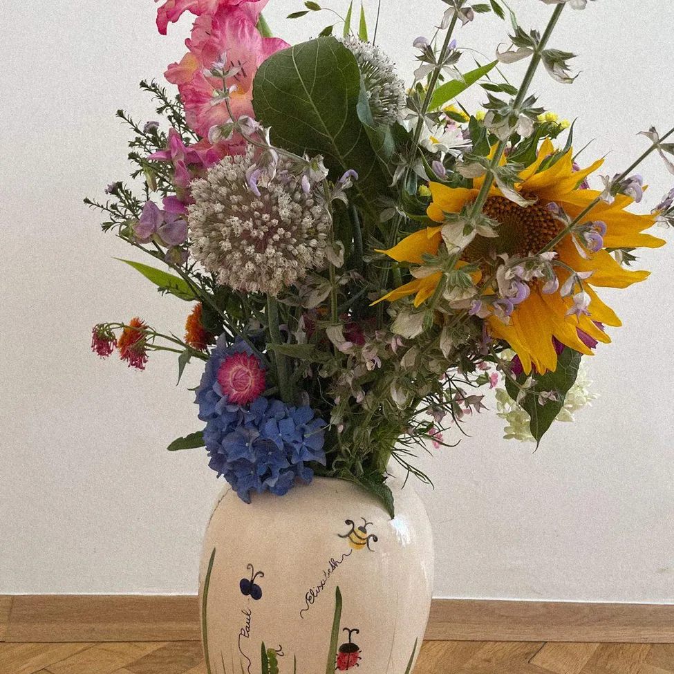 Eine weiße Keramikvase mit gemalten Marienkäfern und Text steht auf einem Holzboden und hält einen bunten Blumenstrauß mit rosa, blauen und gelben Blüten.
