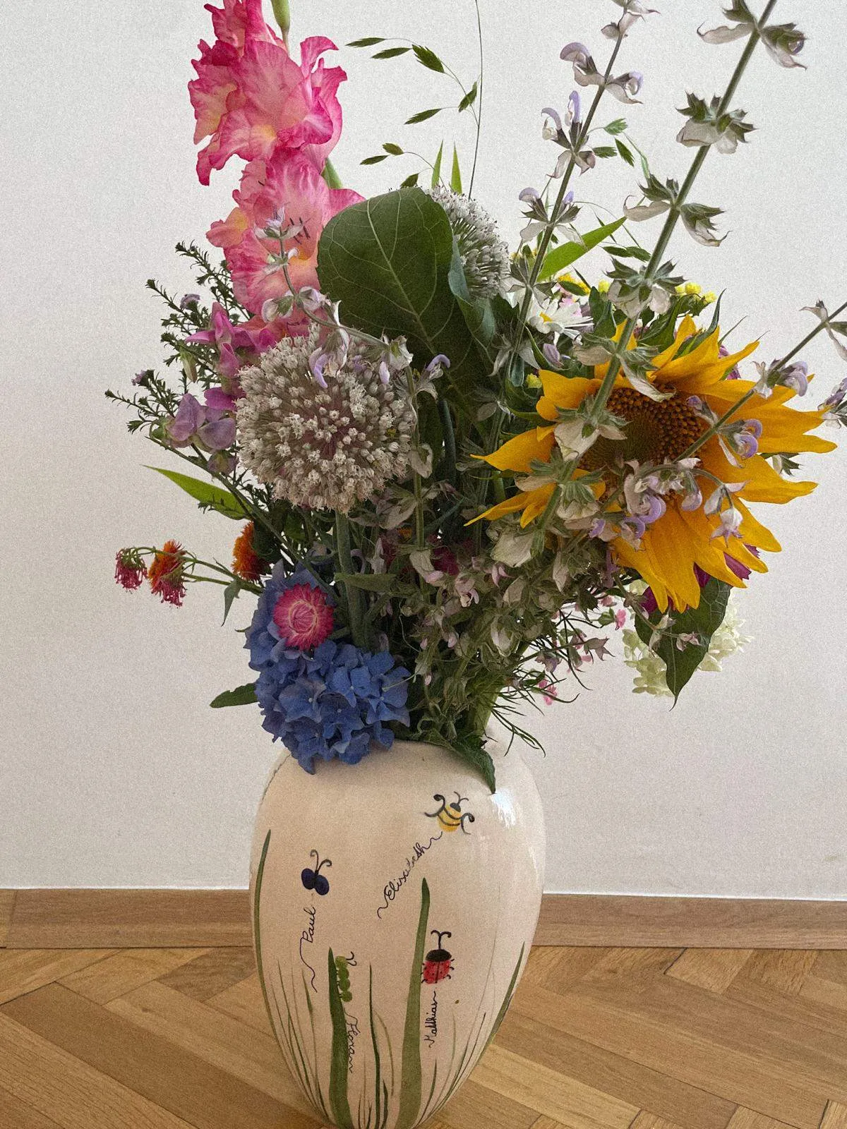 Eine weiße Keramikvase mit gemalten Marienkäfern und Text steht auf einem Holzboden und hält einen bunten Blumenstrauß mit rosa, blauen und gelben Blüten.