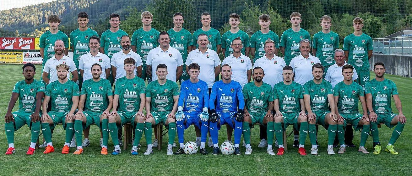 Eine Fußballmannschaft posiert für ein Foto auf einem Feld, mit Spielern in grünen und blauen Trikots, einem Trainer in einem weißen Hemd und zwei Torhütern in Blau.