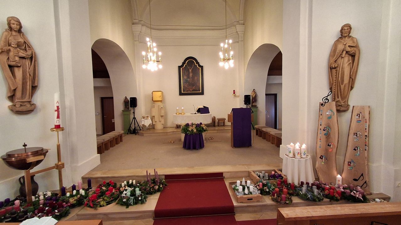 Eine leere Kirche mit einem blumigen Altaraufbau, einem roten Teppich zu den Bänken, zwei Kronleuchtern und einem Bilderrahmen an der Wand.