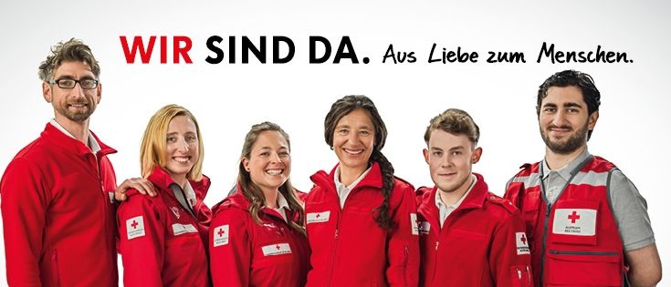 Eine Gruppe von Freiwilligen in roten Uniformen mit weißen Kreuzen auf der Brust lächelt für ein Foto. Der Text über ihnen lautet 'Wir sind da. Aus Liebe zum Menschen.' auf Deutsch.