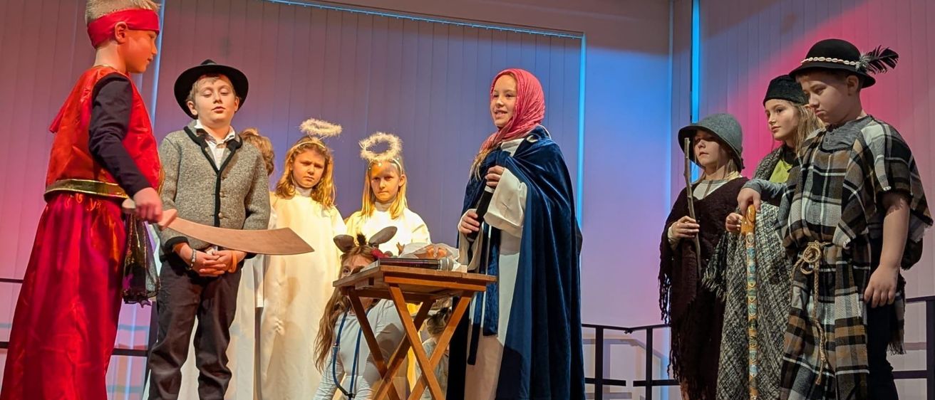 Kinder in verschiedenen Kostümen spielen auf der Bühne mit einer Frau, die ein Buch hält. Einige halten Requisiten, und ein kleiner Tisch ist vor ihnen aufgestellt. Die Bühne hat einen Hintergrund mit vertikalen weißen und blauen Streifen.