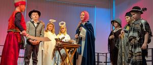 Kinder in verschiedenen Kostümen spielen auf der Bühne mit einer Frau, die ein Buch hält. Einige halten Requisiten, und ein kleiner Tisch ist vor ihnen aufgestellt. Die Bühne hat einen Hintergrund mit vertikalen weißen und blauen Streifen.