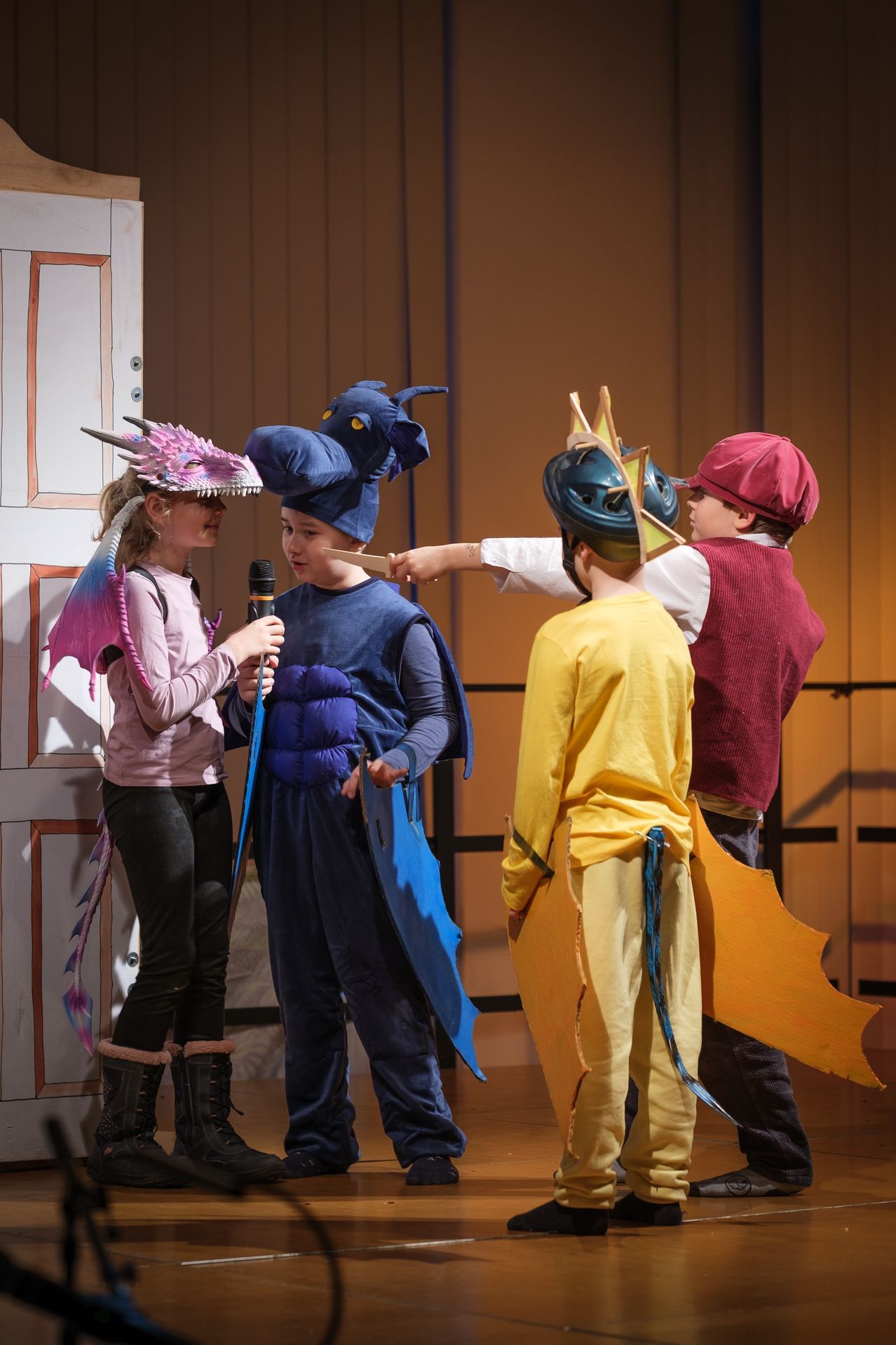 Four children dressed in dragon costumes are on a stage, one holding a microphone, another gesturing with a stick, and the others facing the audience.