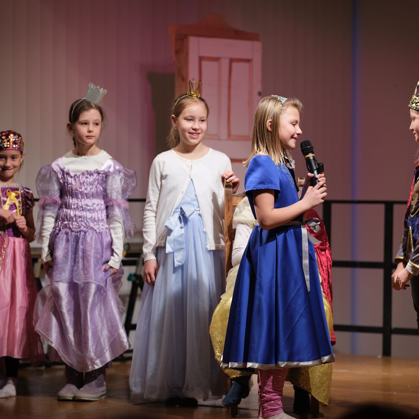Five girls in costumes on a stage, one holding a microphone, another with a crown, and three smiling at the camera.