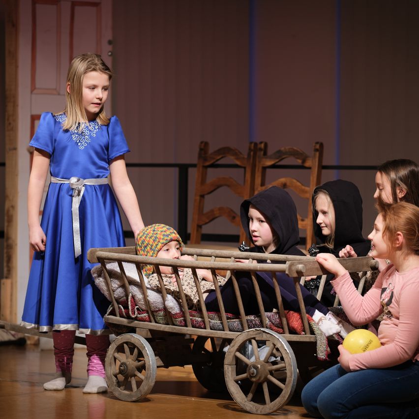 A young girl in a blue dress stands by a wooden cart with a baby inside. Other children sit around the cart, engaging with the baby.
