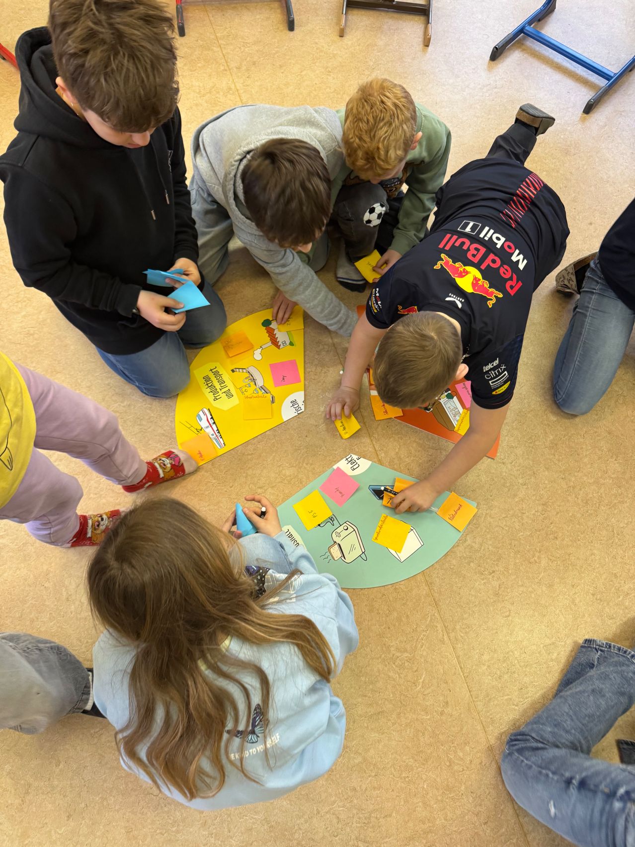 Eine Gruppe von Kindern sitzt auf dem Boden, vermutlich in einem Klassenzimmer, und scheint eine Aktivität durchzuführen. Einige von ihnen halten Klebezettel, während andere auf den Boden schreiben.