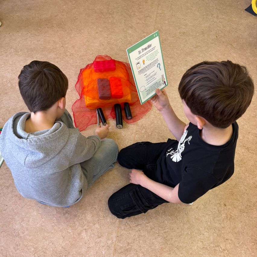 Zwei junge Jungen sitzen auf dem Boden und beschäftigen sich mit einer Aktivität. Ein Junge hält ein grünes Büchlein, während der andere schwarze und orangefarbene Objekte manipuliert.