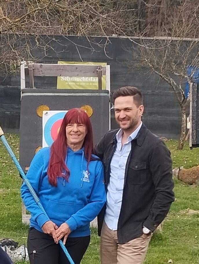 Ein Mann und eine Frau stehen lächelnd vor einem Ziel für eine Bogenschießanlage. Die Frau hält einen Bogen, während der Mann neben ihr steht, beide lässig gekleidet.