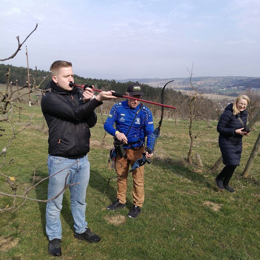 Drei Personen sind in einem Feld mit Bögen, eine mit einem Bogen, eine mit einer Kamera und eine, die in ein Horn bläst.