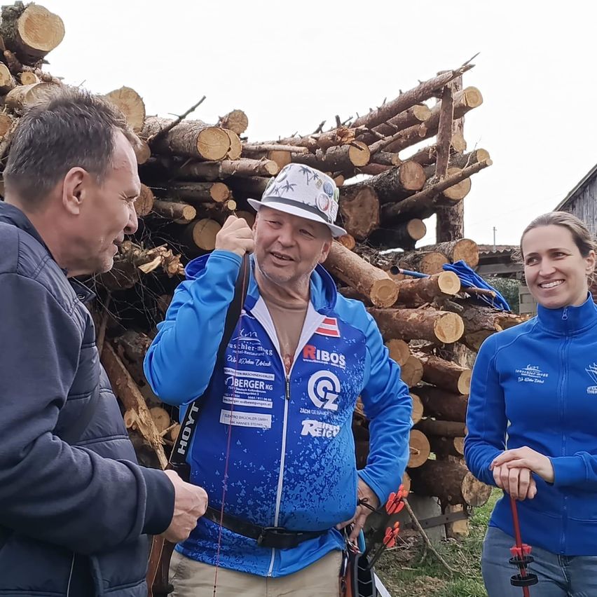 Drei Personen, zwei Männer und eine Frau, stehen vor einem Stapel Holzscheite. Sie tragen alle blaue Sportjacken, wobei der Mann in der Mitte einen Hut trägt und einen Bogen hält.