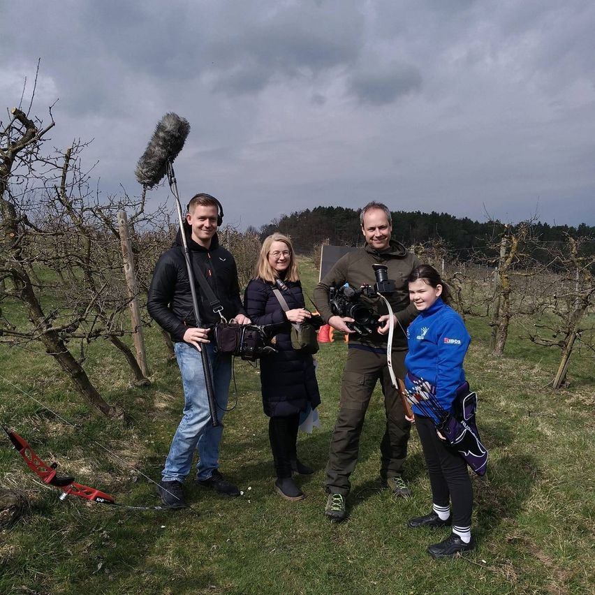 Vier Personen stehen in einem Obstgarten. Eine Person hält einen Bogen. Eine weitere Person hält eine Kamera. Sie alle lächeln.