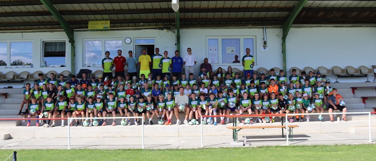 Eine große Gruppe von Kindern und Erwachsenen in grünen, gelben und roten Trikots posiert für ein Foto auf einem Fußballfeld, mit einer Bank und einem Torpfosten im Vordergrund.