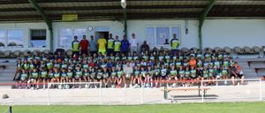 Eine große Gruppe von Kindern und Erwachsenen in grünen, gelben und roten Trikots posiert für ein Foto auf einem Fußballfeld, mit einer Bank und einem Torpfosten im Vordergrund.