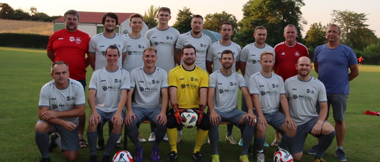 Eine Fußballmannschaft in grauen und weißen Trikots posiert für ein Foto auf einem Rasenfeld mit Bäumen im Hintergrund. Ein Mann in einem gelben Trikot hält einen Fußball.