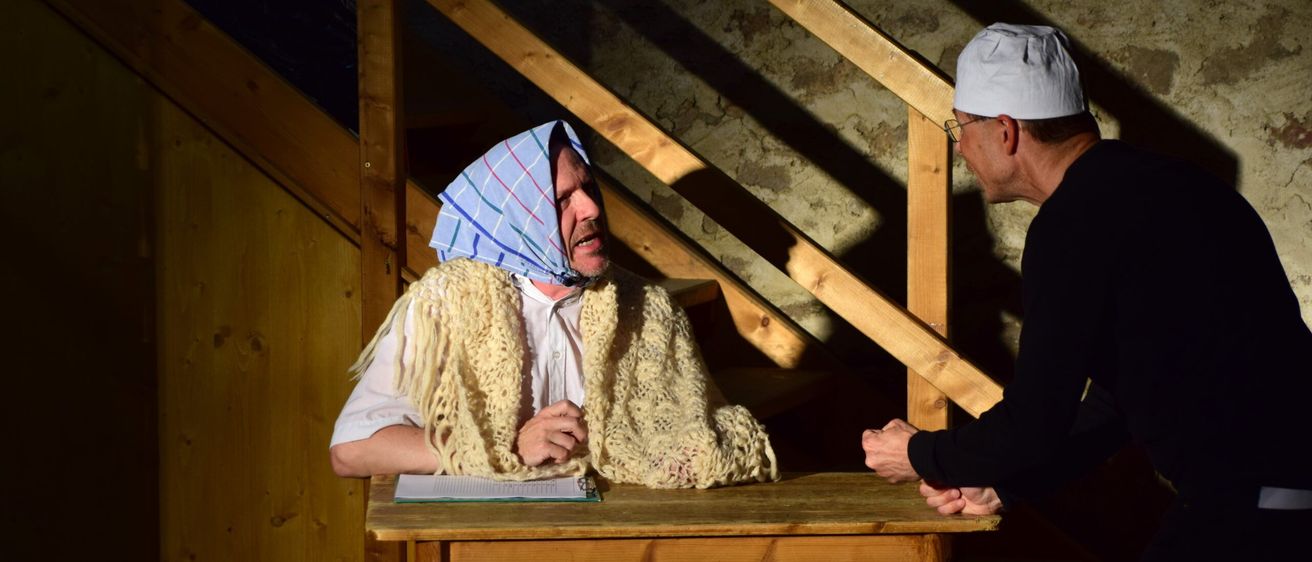 Ein Mann in traditioneller Kleidung sitzt mit einem Buch an einem Holztisch, während ein anderer Mann mit einer weißen Haube zusieht.