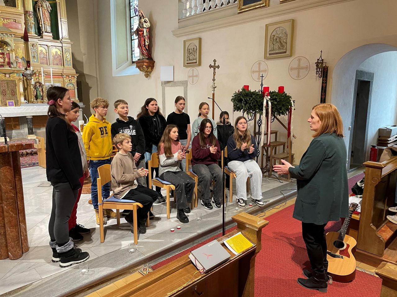 Ein Kinderchor singt in einer Kirche, wobei eine Frau zu ihnen spricht. Der Chor sitzt auf Stühlen auf einer Bühne. Hinter ihnen steht eine Frau mit einem Mikrofon.