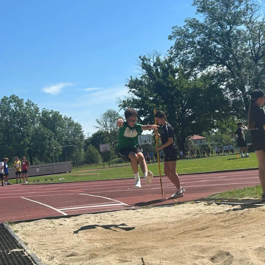 Ein Junge springt über eine Hürde bei einer Leichtathletikveranstaltung, während eine Frau seinen Sprung misst.