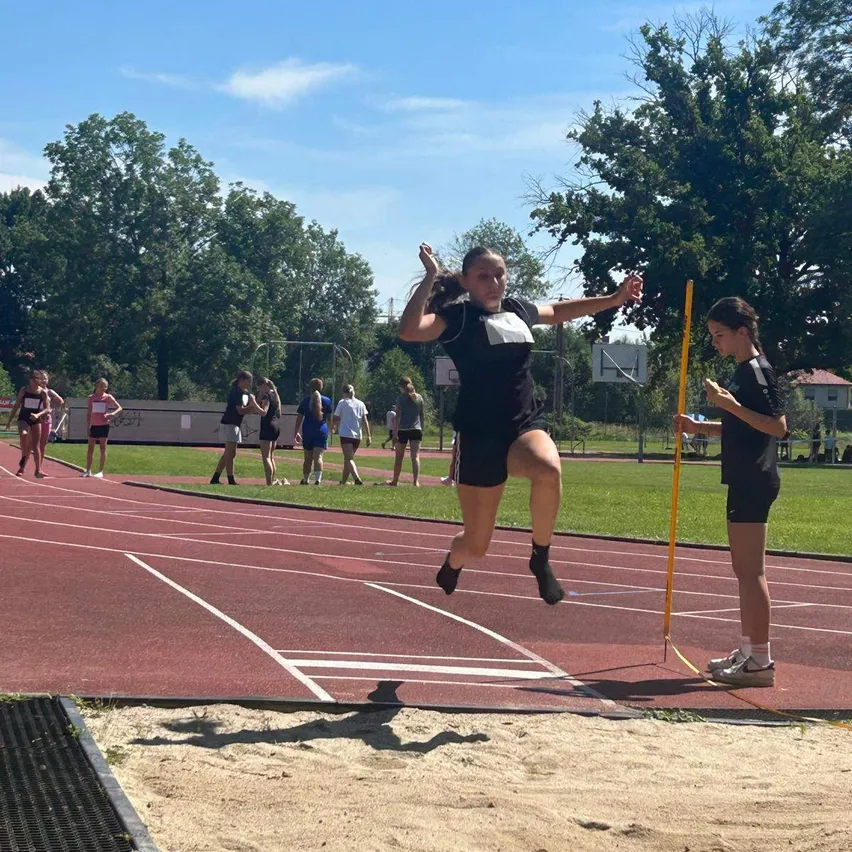 Eine junge Frau führt einen Weitsprung auf einer Outdoor-Laufbahn durch, während andere zusehen und ihren Sprung messen.